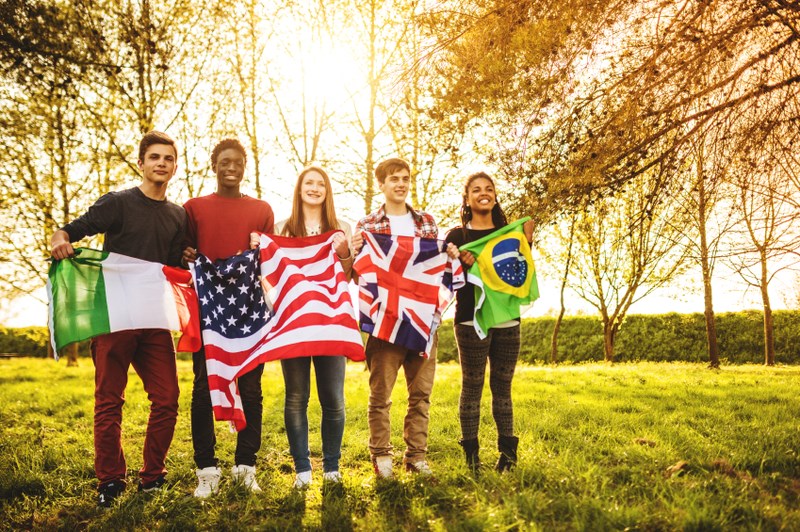 5 male and female students standing outside in a wooded area holding flags from 4 different countries in front of them. 