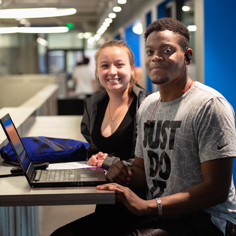 Two students sitting together with a laptop smiling at the camera