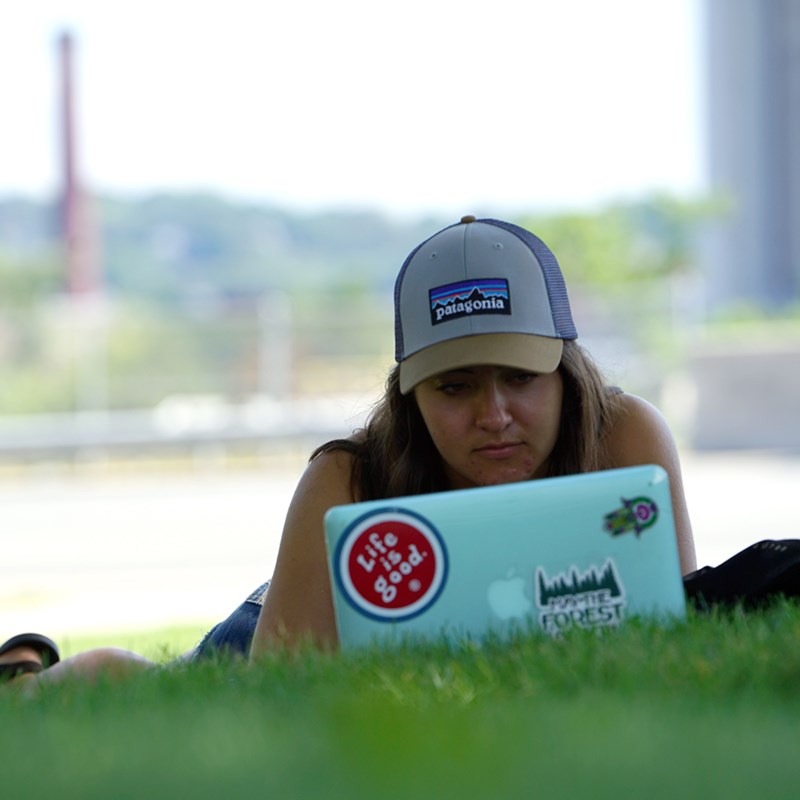 A female student wearing a hat lays on green grass and studies on a laptop, behind them you can see Fox Hall