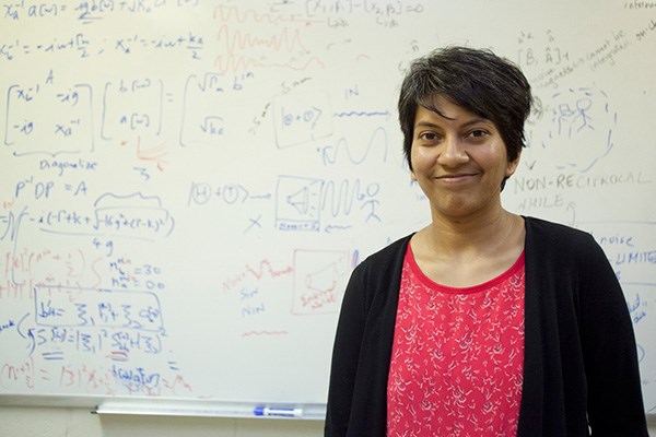 UMass Lowell physics professor Archana Kamal stands in her office in front of a whiteboard covered with formulas and notes.