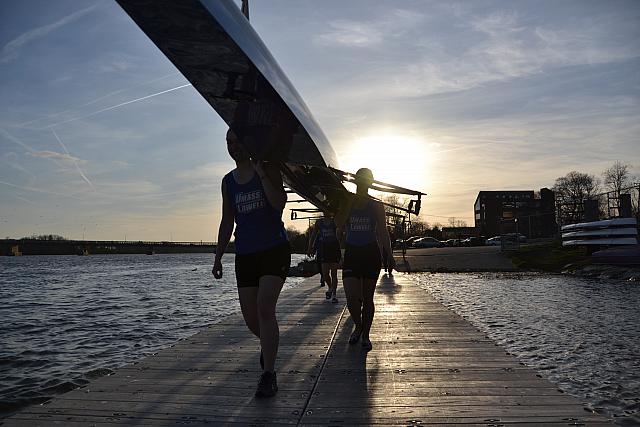 UMass Lowell rowers walking with shell (boat) overhead on a dock during sunset.