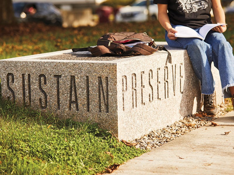 Sustain/Preserve chiseled on stone planter where young person is sitting.