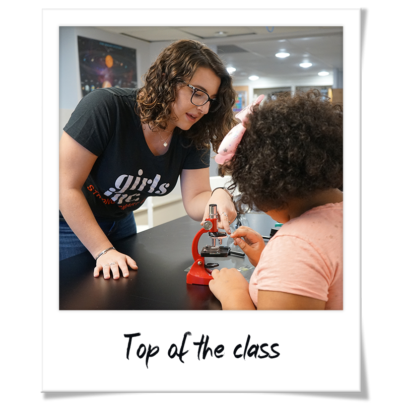 "Polaroid" photo of Sydney Fagundes showing a girl how to use a microscope at a summer program though Girls Inc. of Greater Lowell - handwritten frame reads "Top of the class"
