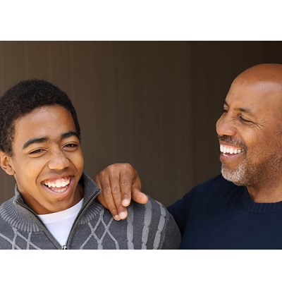 A teenage African-american boy laughing along with his African-american father who has his hand on the boy's shoulder.