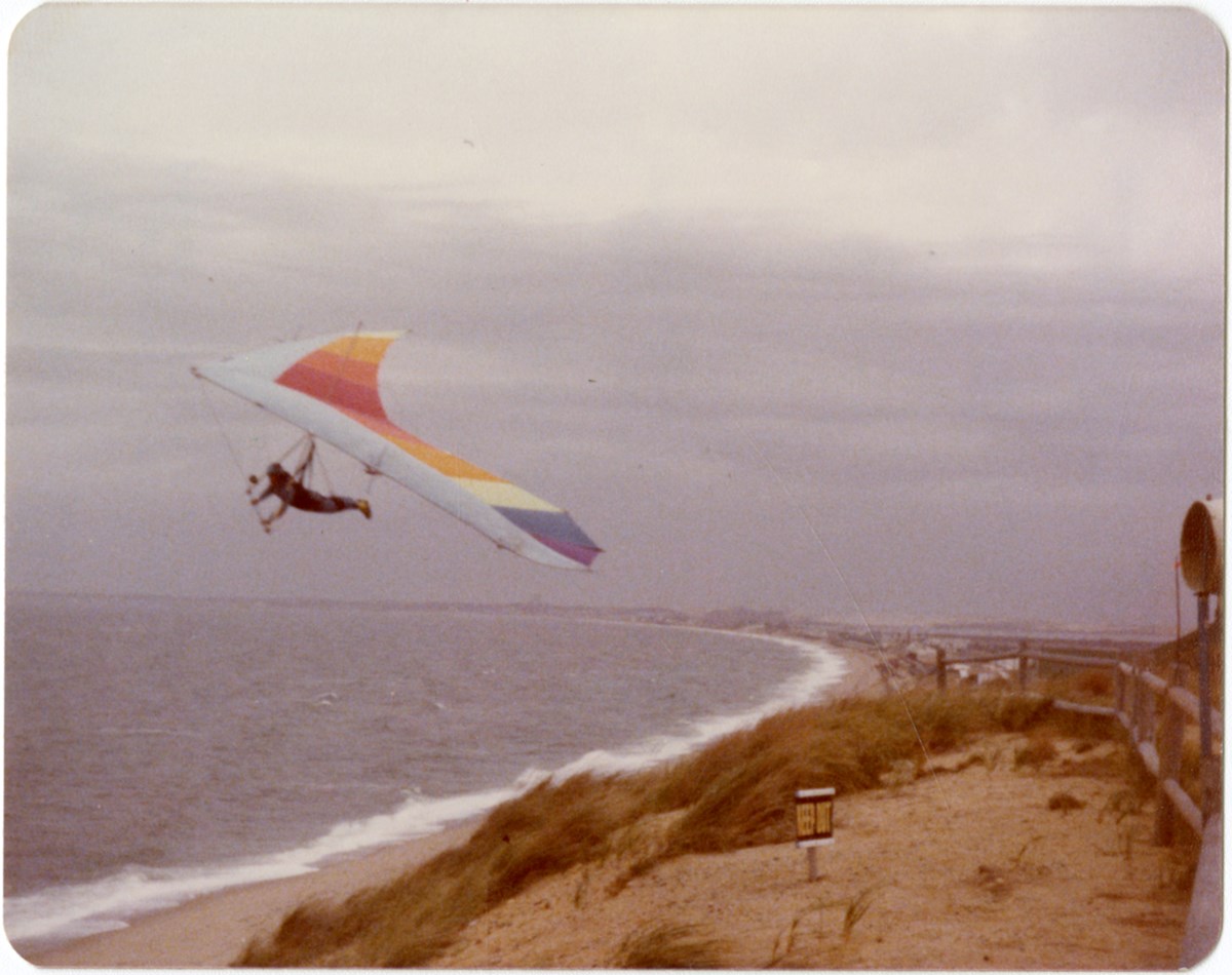 An old color photo of a hang glider soaring over a beach