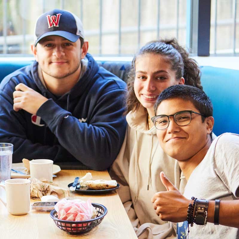 Three students sit at a dining hall table and one students shows a thumbs up