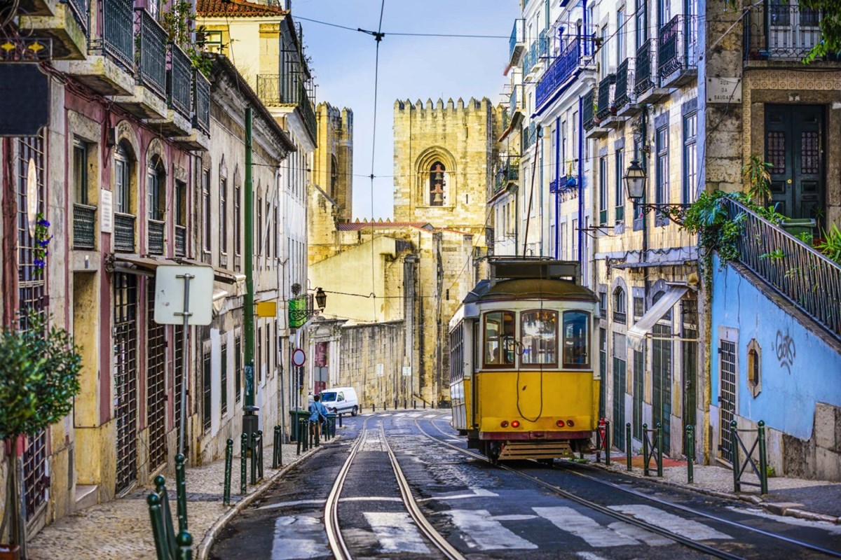 A street in Old town in Lisbon, Portugal with a yellow Tram in the foreground and a cathedral looming in the background and buildings on both sides.