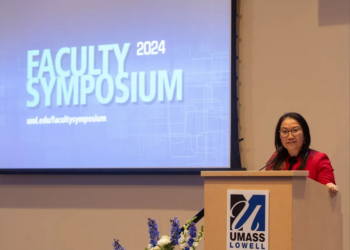 UMass Lowell Faculty member giving a speech with 2024 Faculty Symposium – uml.edu/facultysymposium on the screen.