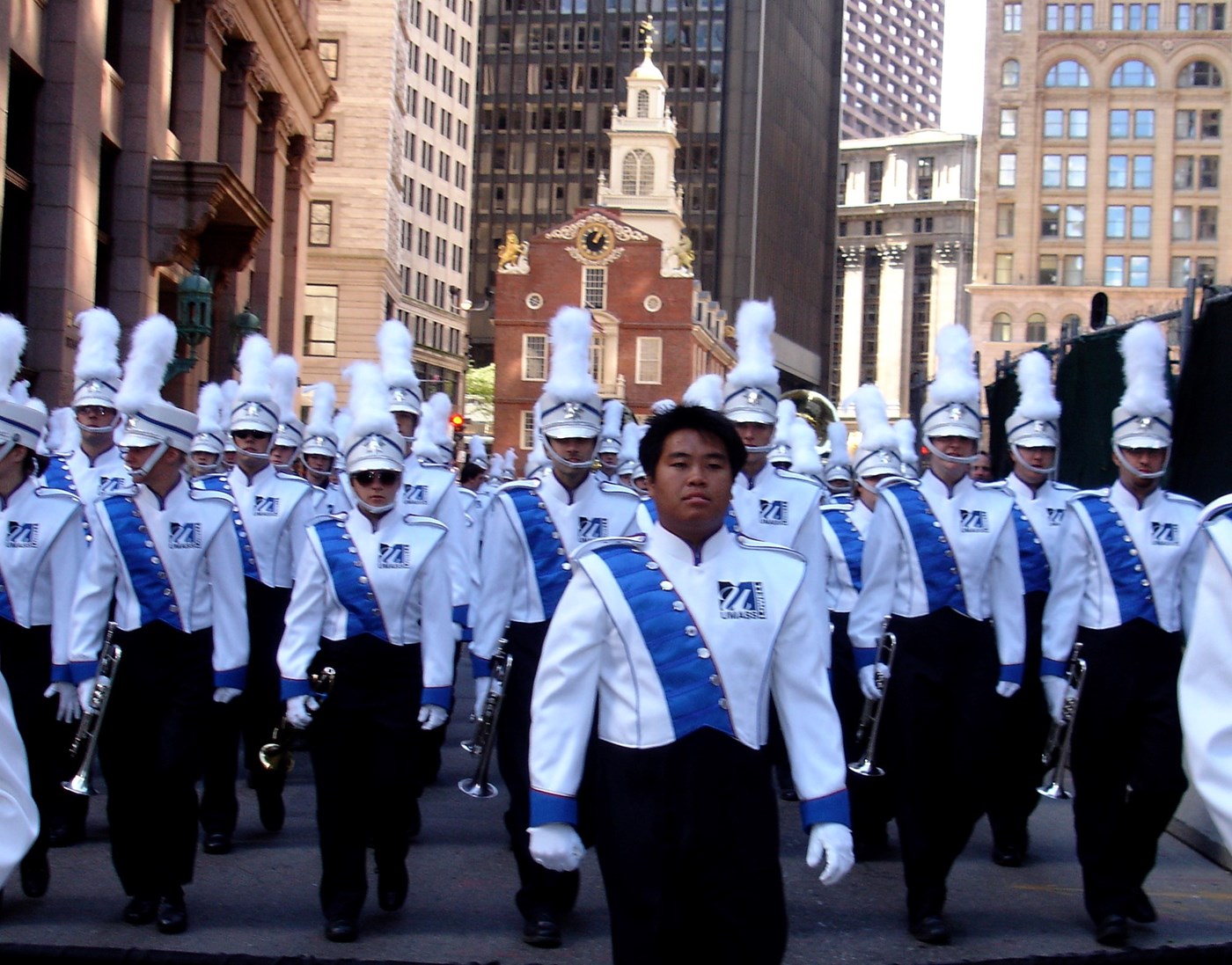 The UMass Lowell Marching Band's iconic photo shows the band in parade formation in Boston, marching in their white uniform tops with royal blue sash, lead by their Asian student field conductor.