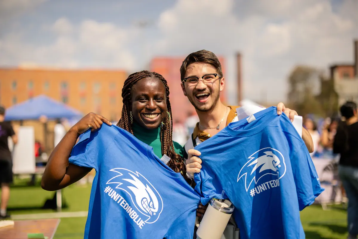 Two students holding up blue T-shirts with River Hawk logo and #unitedinblue underneath