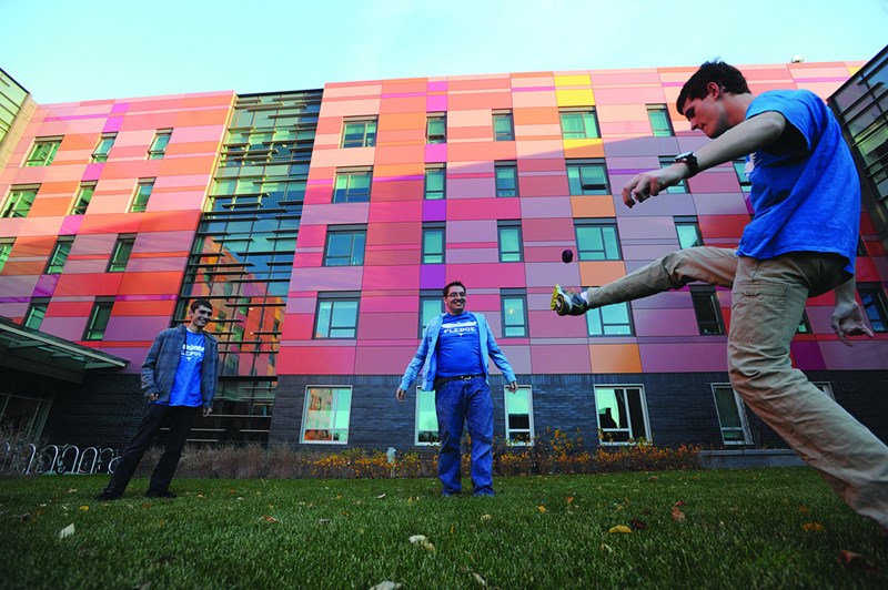 Three male students play with a hacky sack on the University Suites quad