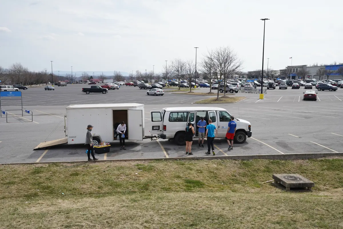 A van and trailer are stopped in a large parking lot with a few people standing around.