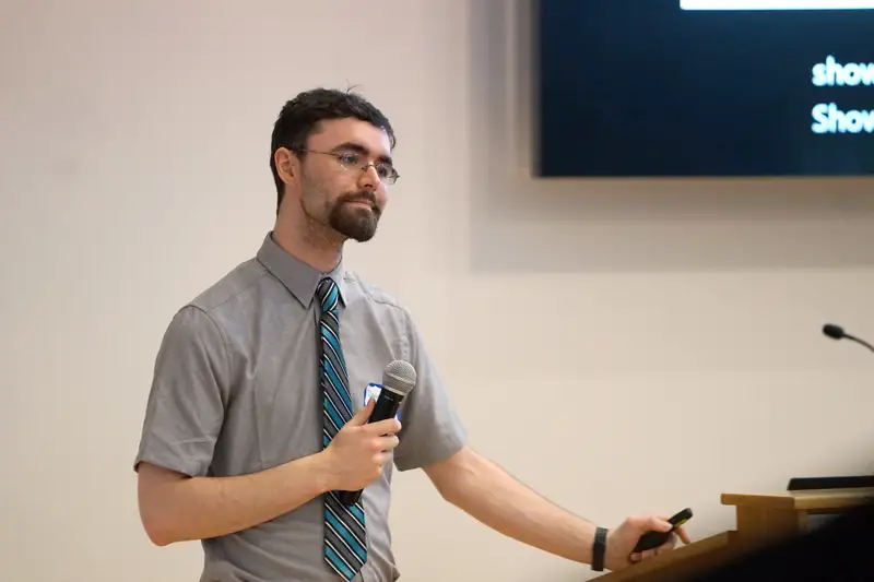 Dawson Durgin, team leader for Votecational, dressed in a shirt and tie, holding a microphone and looking at the (unseen) audience.