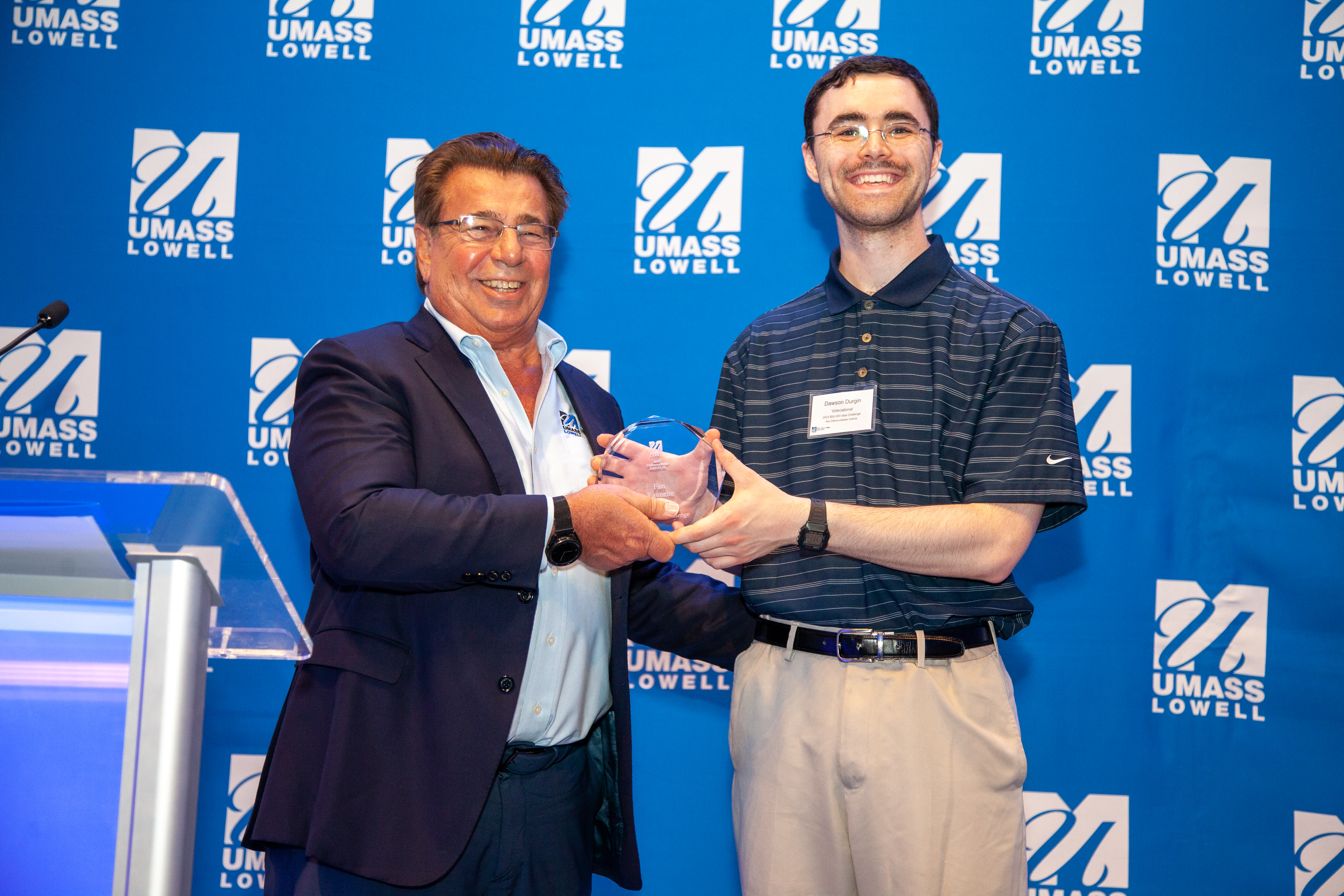 Brian Rist and Dawson Durgin, team leader for Votecational smiling and holding the Fan Favorite award in front of a UMass Lowell blue background.