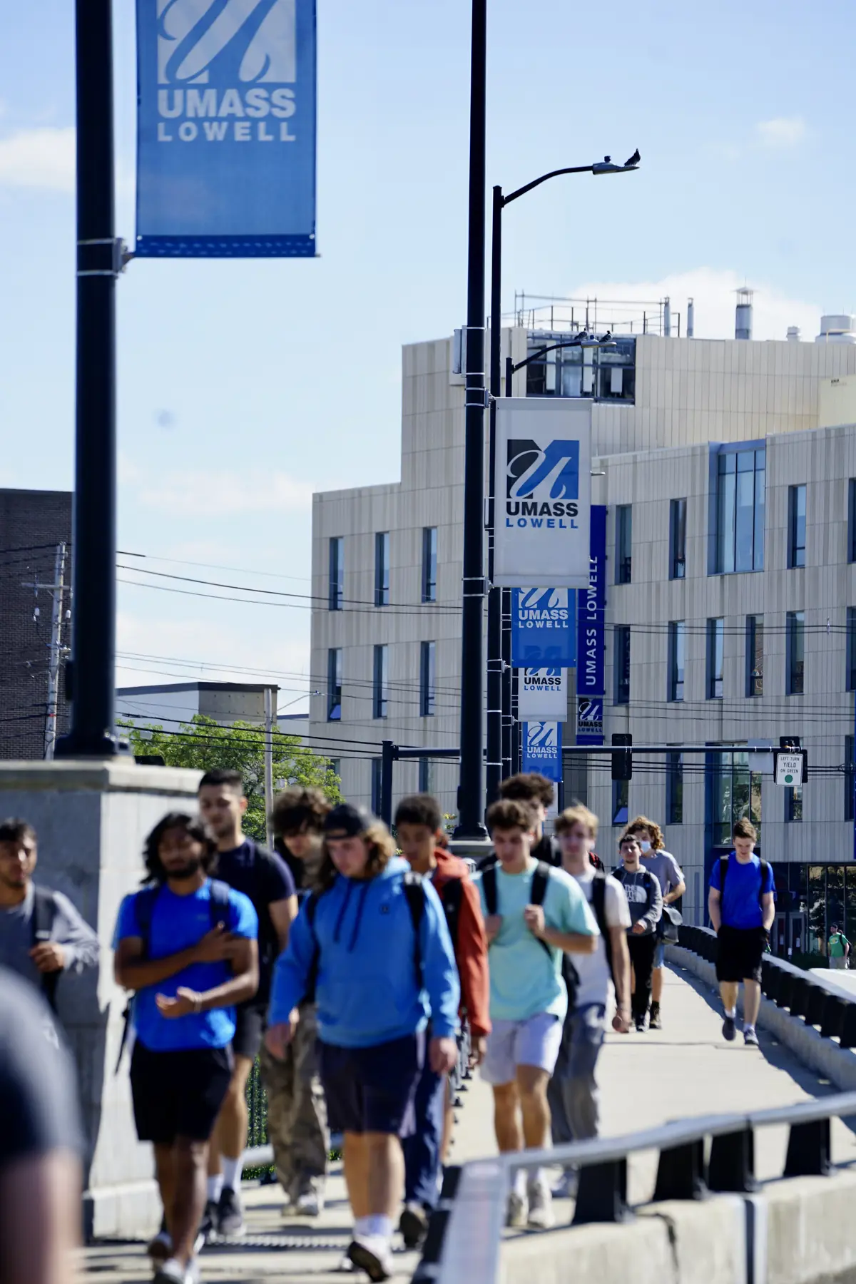 A bevy of students walks along a sidewalk beneath UMass Lowell banners hanging from lampposts