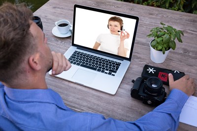 An image of two colleagues collaborating on (or discussing) a work-in-progress through video conference call on a laptop. Man sitting at table looking at laptop computer with woman wearing a headset and talking.
