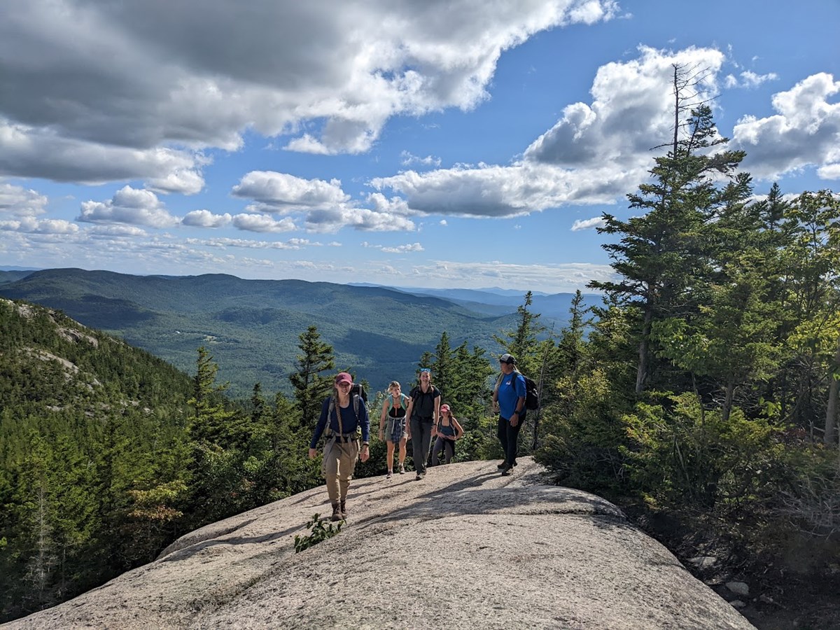 5 people walk on an open rock ledge with surrounding mountain views with blue sky and clouds