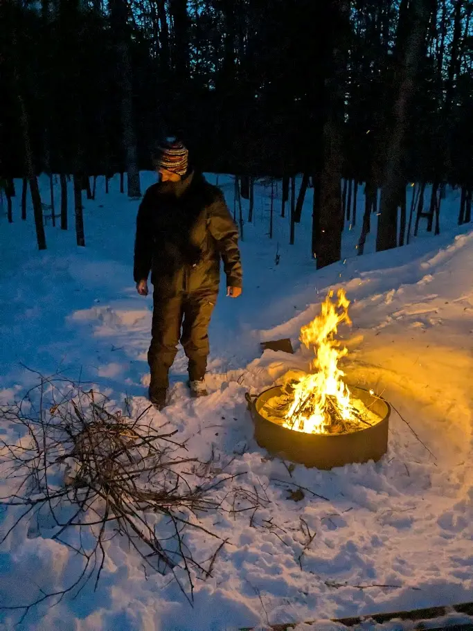 A bright campfire lights the snow in the dusk as a person reaaches for branches.