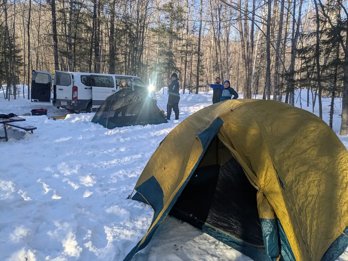 A couple tents are set up in the snow with a few smiling people and a van, with the sun shining.