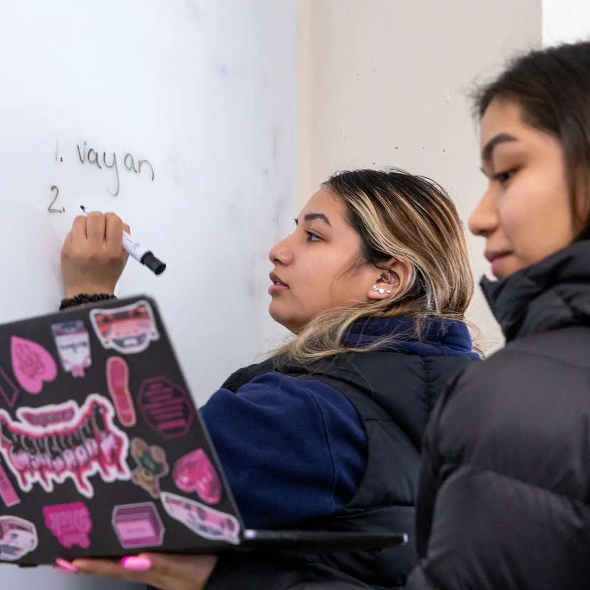 A student writes the word "vanyan" on a white board while another student holds up a laptop.