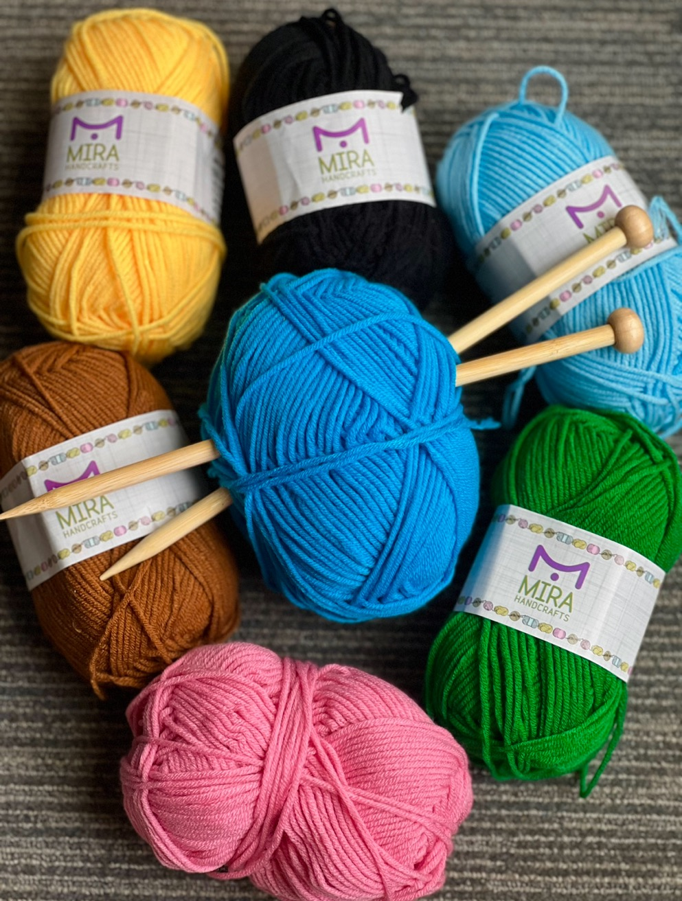 Bundles of colored yarn with the label "Mira Handcrafts" around them. 2 tan knitting needles going through the blue yarn at the center.