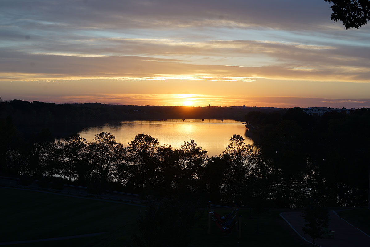 Sunset over a calm river with silhouetted trees, colorful sky reflections, and a park with hammocks in the foreground