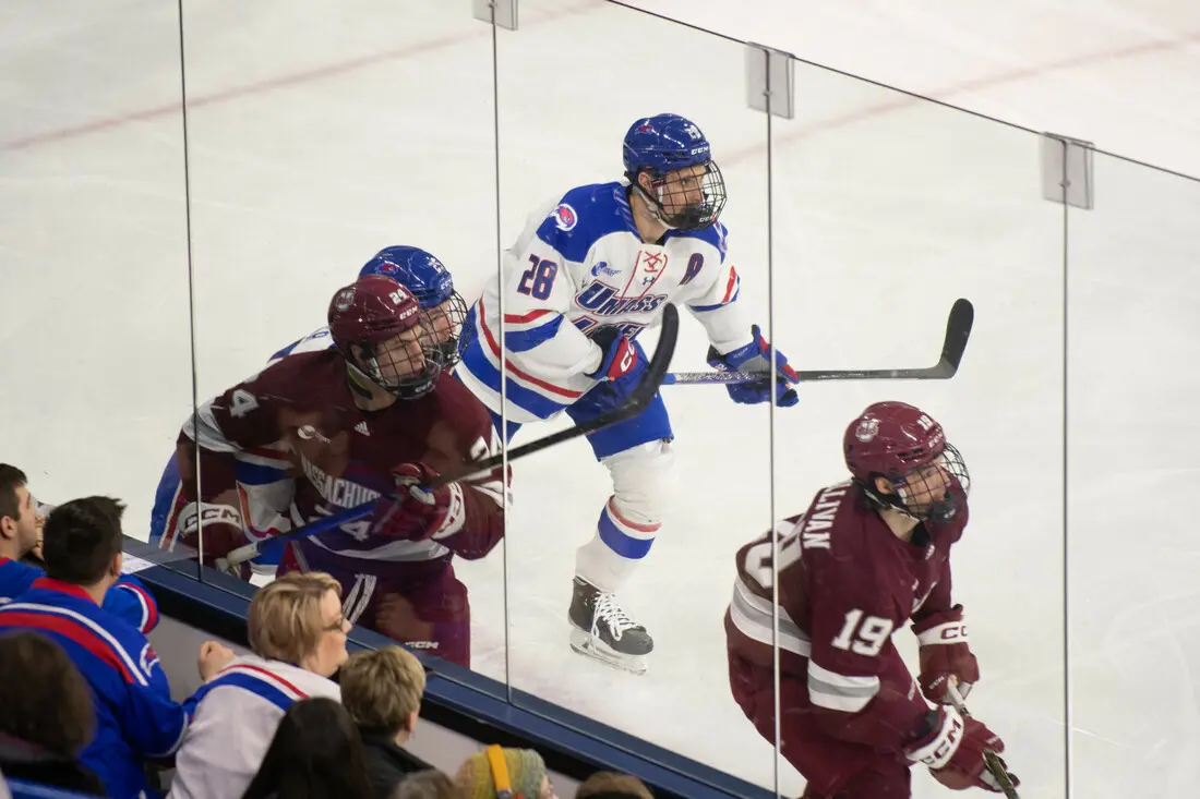 Hockey players compete on the ice, with one in a white jersey and two in maroon jerseys near the glass