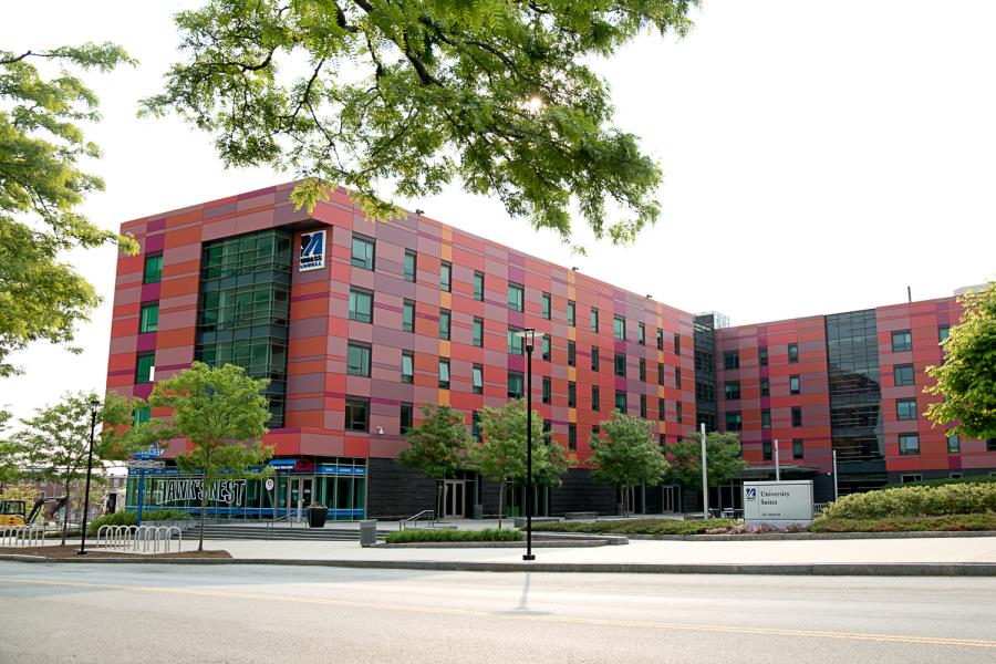 Pink and purple brick five-story, L-shaped building, with University Suites sign on lawn out front