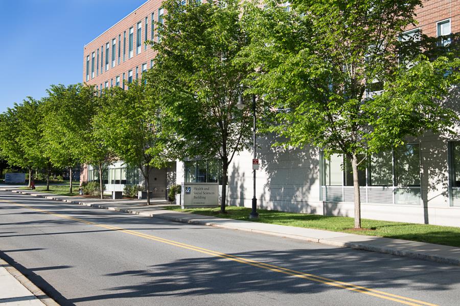 Sign for Health and Social Sciences Building along tree-lined sidewalk in front of modern brick classroom building