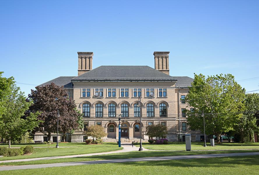 Grand, dual chimneyed tan brick building with three archways at entrance and arched windows along second of three stories