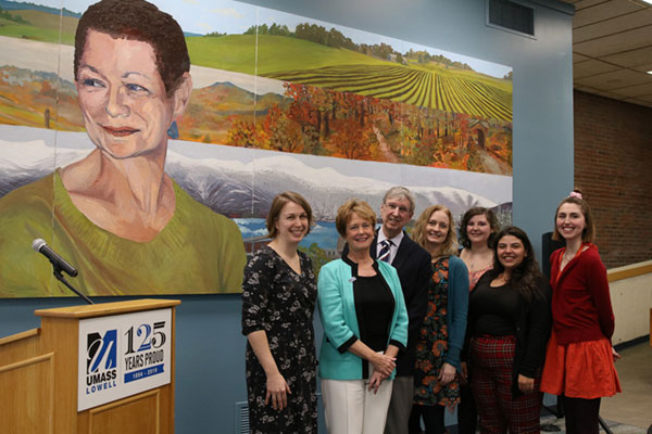 Group of people including Chancellor Jacquie Moloney stand in front of Nina Coppens mural in O'Leary Library.