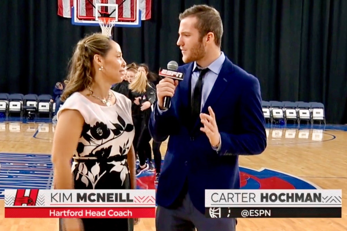 A person in a suit holding a microphone interviews a person in a white and black top on a basketball court.