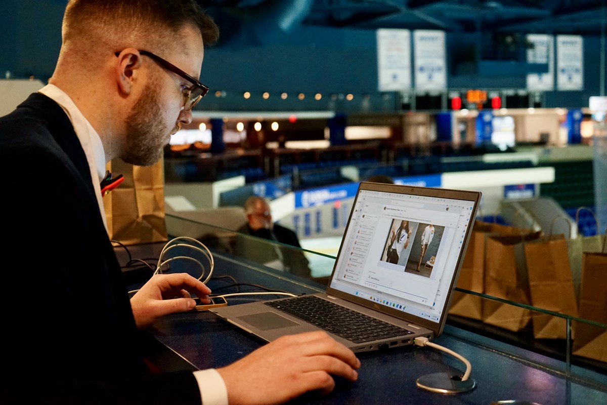 A person in a suit and glasses works on a laptop at a table in an arena.