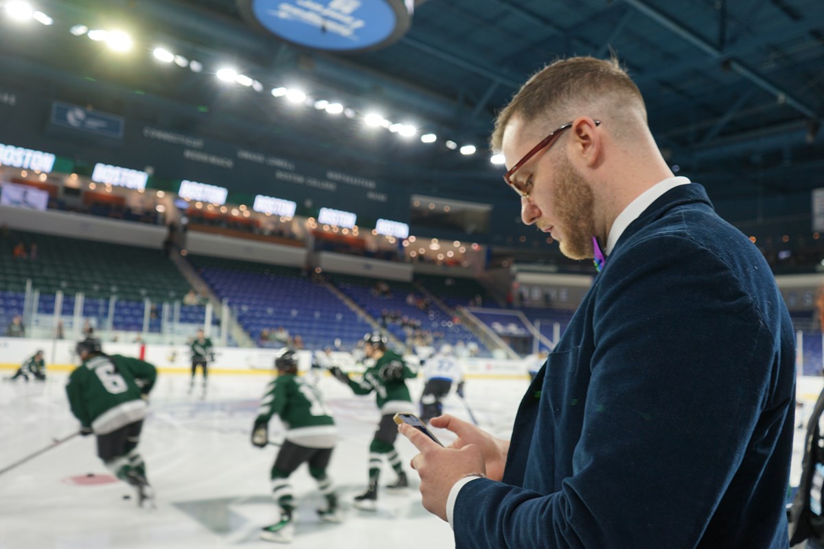 A person looks at their phone while standing next to an ice rink where hockey players are skating.